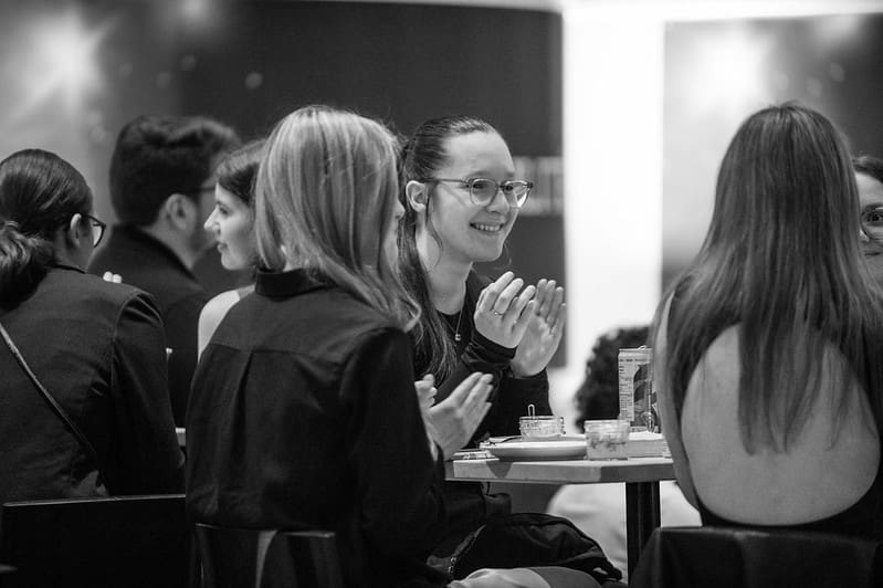 Un groupe de personnes est assis autour d'une table ; certaines sourient et applaudissent. Le cadre semble correspondre à un événement social ou professionnel. La photo est en noir et blanc.