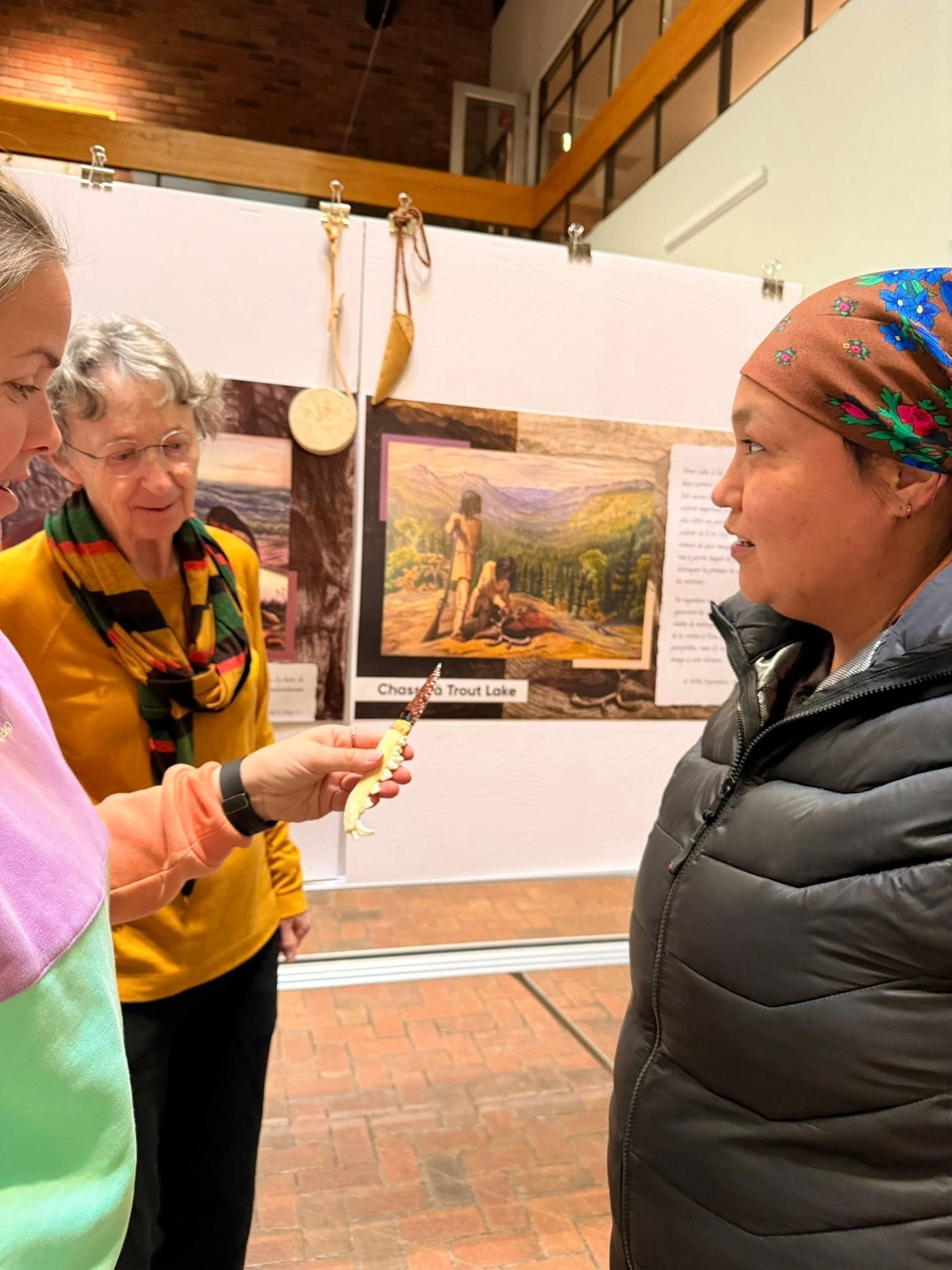Trois femmes discutent à l'intérieur, près d'une vitrine. L'une d'elles tient un petit objet orné de perles, tandis que les autres l'observent. On aperçoit des œuvres d'art et des textes explicatifs à l'arrière-plan.