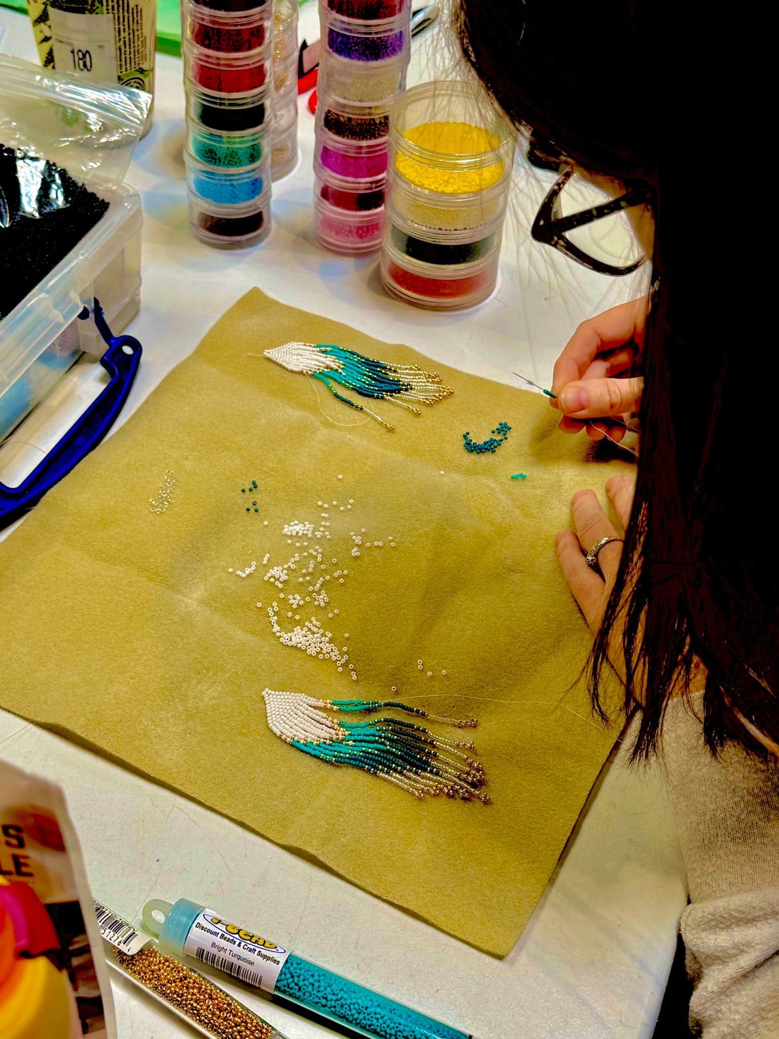 Une personne s'affaire à confectionner des boucles d'oreilles en perles de couleur bleu sarcelle, blanches et dorées. Plusieurs petits récipients remplis de perles colorées sont disposés sur la table à proximité.