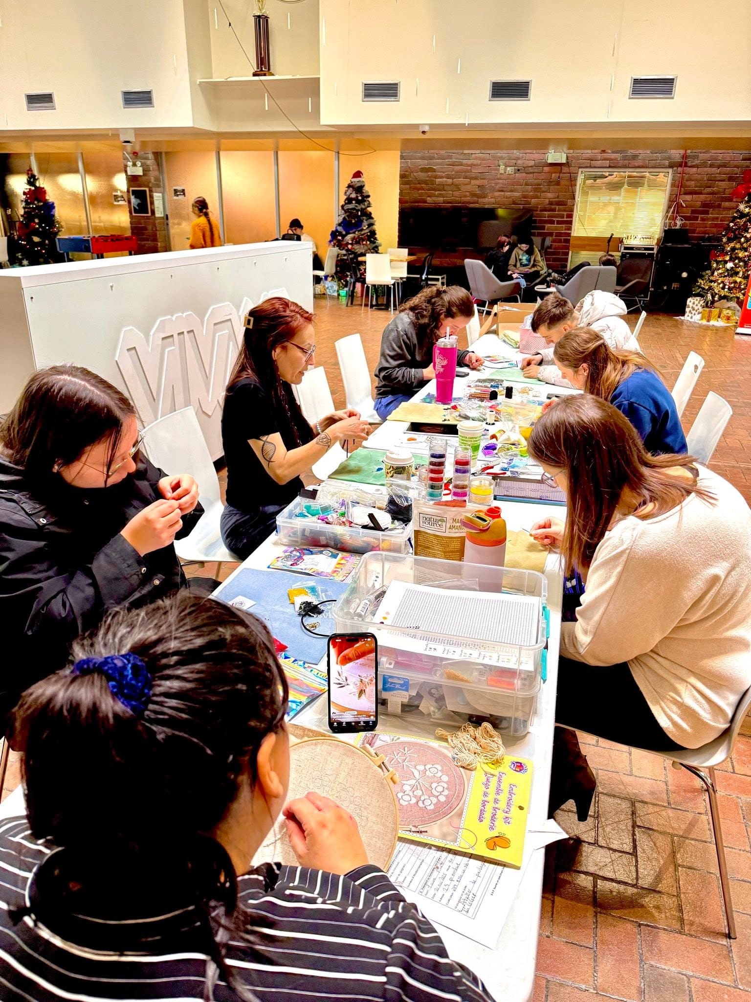 Un groupe de personnes est assis autour d'une table et s'adonne à des activités manuelles dans une pièce bien éclairée, décorée de sapins de Noël et de décorations de fin d'année. Des fournitures artistiques et des kits de loisirs créatifs sont éparpillés sur la table.