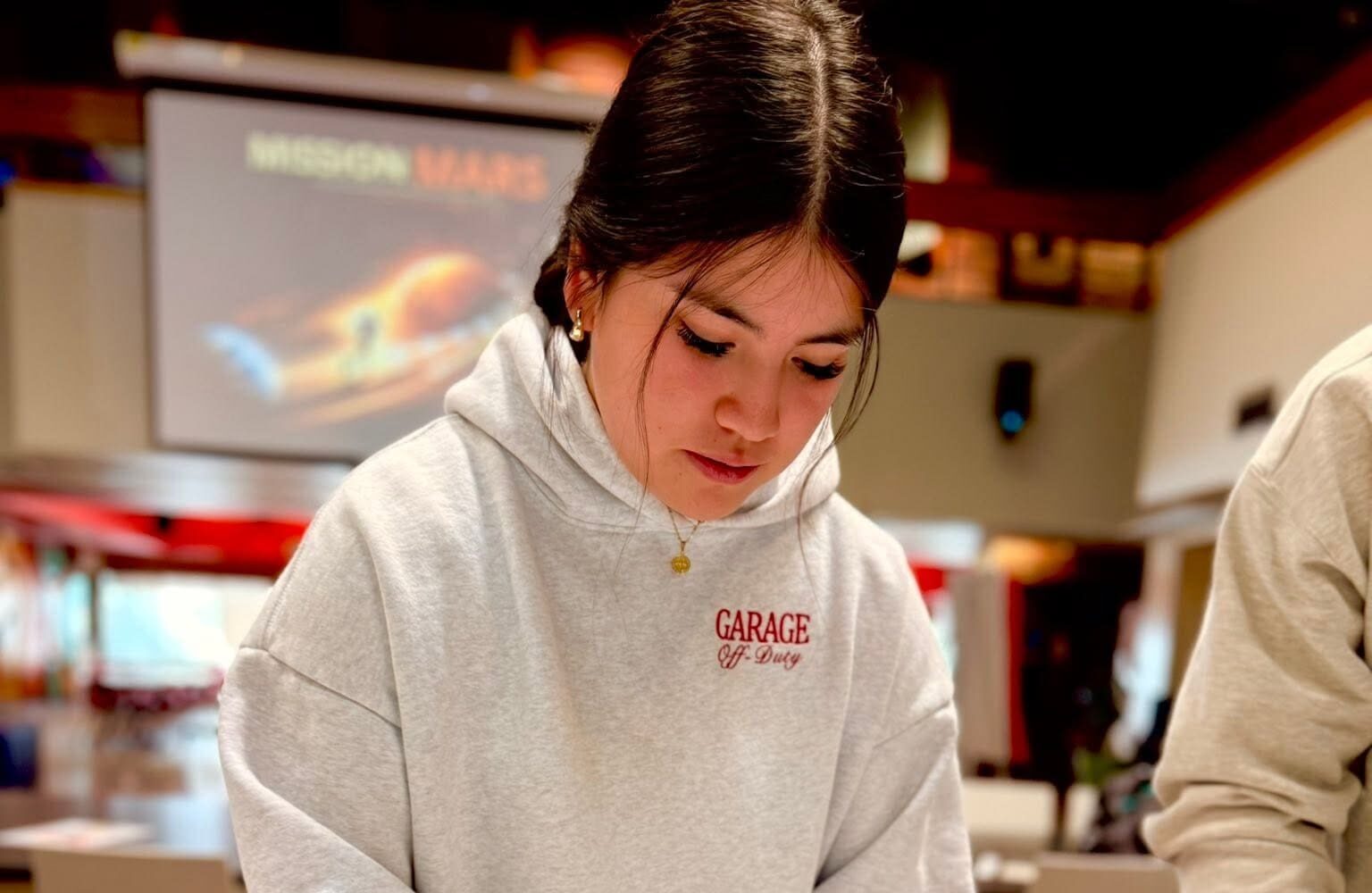 Une jeune femme vêtue d’un sweat à capuche gris clair, sur lequel est inscrit « GARAGE », se concentre sur son travail. Derrière elle, un écran de projection affiche les mots « MISSION MARS » et l’image d’une fusée.