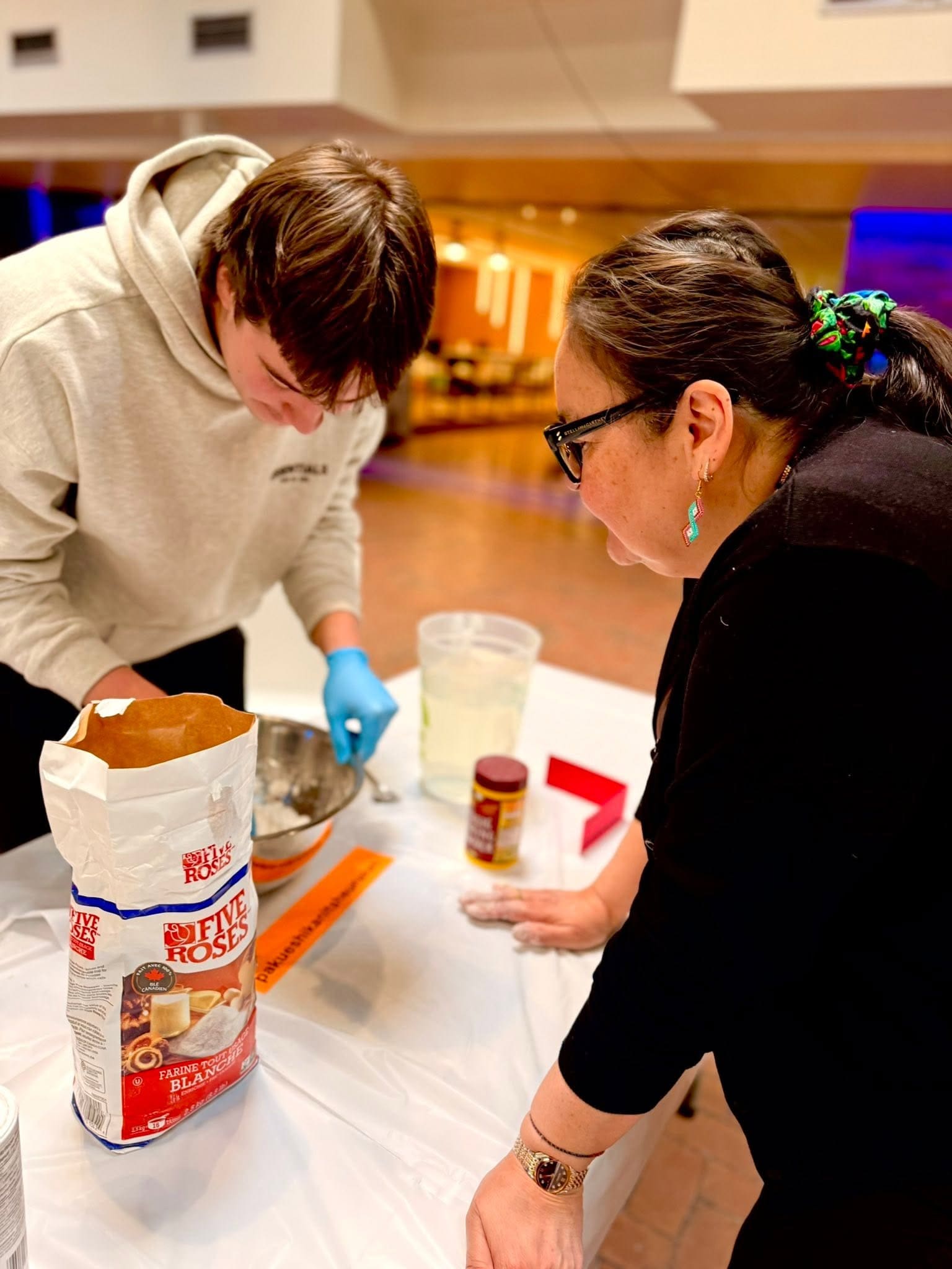 Un adolescent et un adulte préparent des ingrédients sur une table, notamment de la farine, de l'eau et une boîte de conserve. Ils sont en train de cuisiner à l'intérieur.