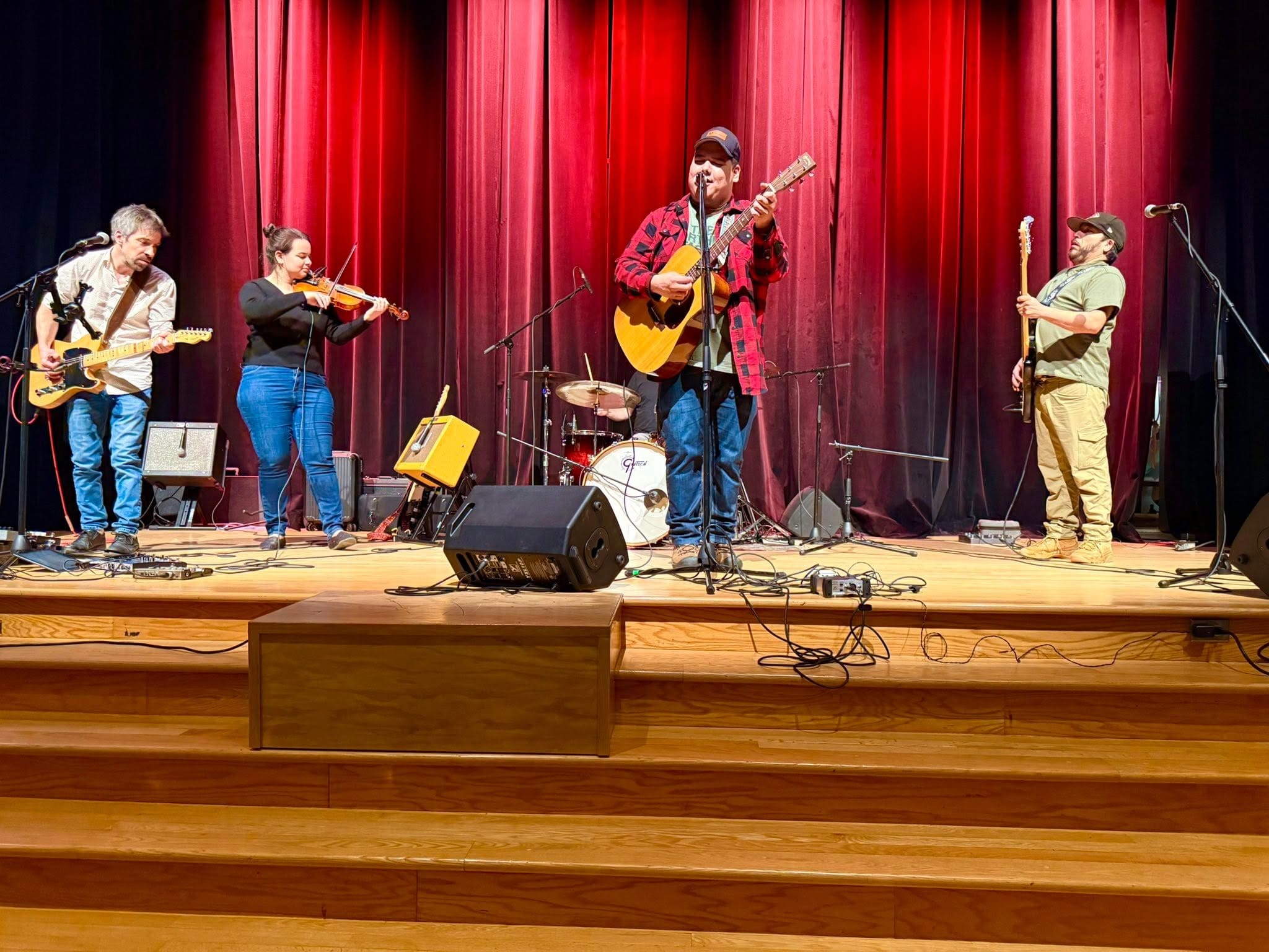 Un groupe se produit sur scène devant des rideaux rouges ; les musiciens jouent de la guitare, du violon, de la batterie et de la basse. Des microphones et des amplificateurs sont installés, et un éclairage chaleureux met les musiciens en valeur.