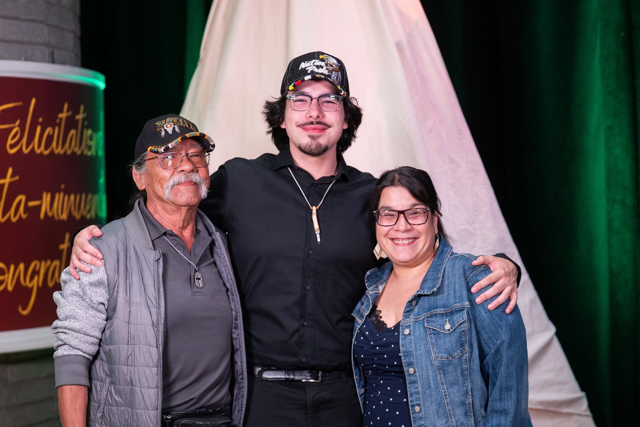 Trois personnes posent ensemble, souriantes, les bras autour les unes des autres. Le groupe se tient devant un tipi blanc, sur fond vert. Les deux hommes portent des casquettes, tandis que la femme porte des lunettes et une veste en jean.
