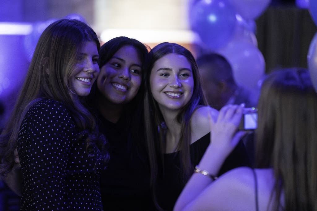Trois jeunes femmes prennent la pose et sourient pour une photo lors d'un événement, tandis qu'une autre personne, au premier plan, les prend en photo. L'arrière-plan est décoré de ballons bleus et blancs.