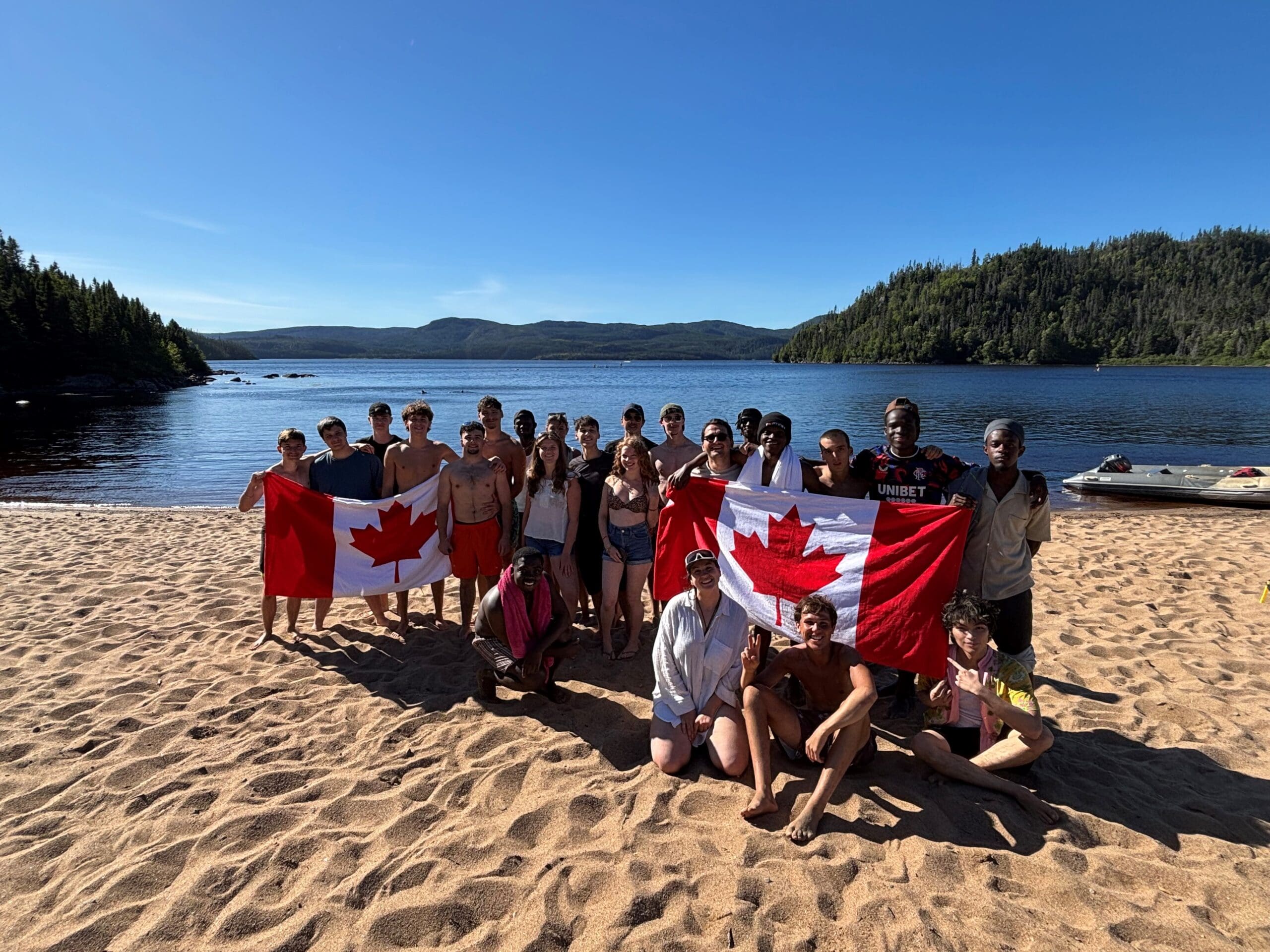 Un groupe de personnes sur une plage de sable au bord d'un lac, brandissant des drapeaux canadiens, avec des arbres et des collines en arrière-plan sous un ciel bleu limpide.