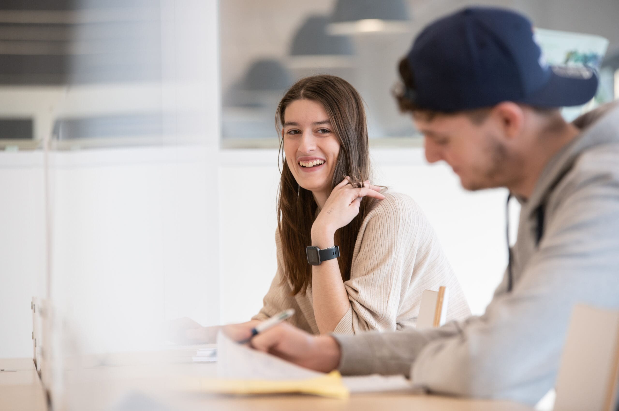 Une jeune femme sourit, assise à un bureau, en regardant un jeune homme qui écrit. Ils semblent se trouver dans une salle de classe ou un espace d'étude lumineux et moderne.