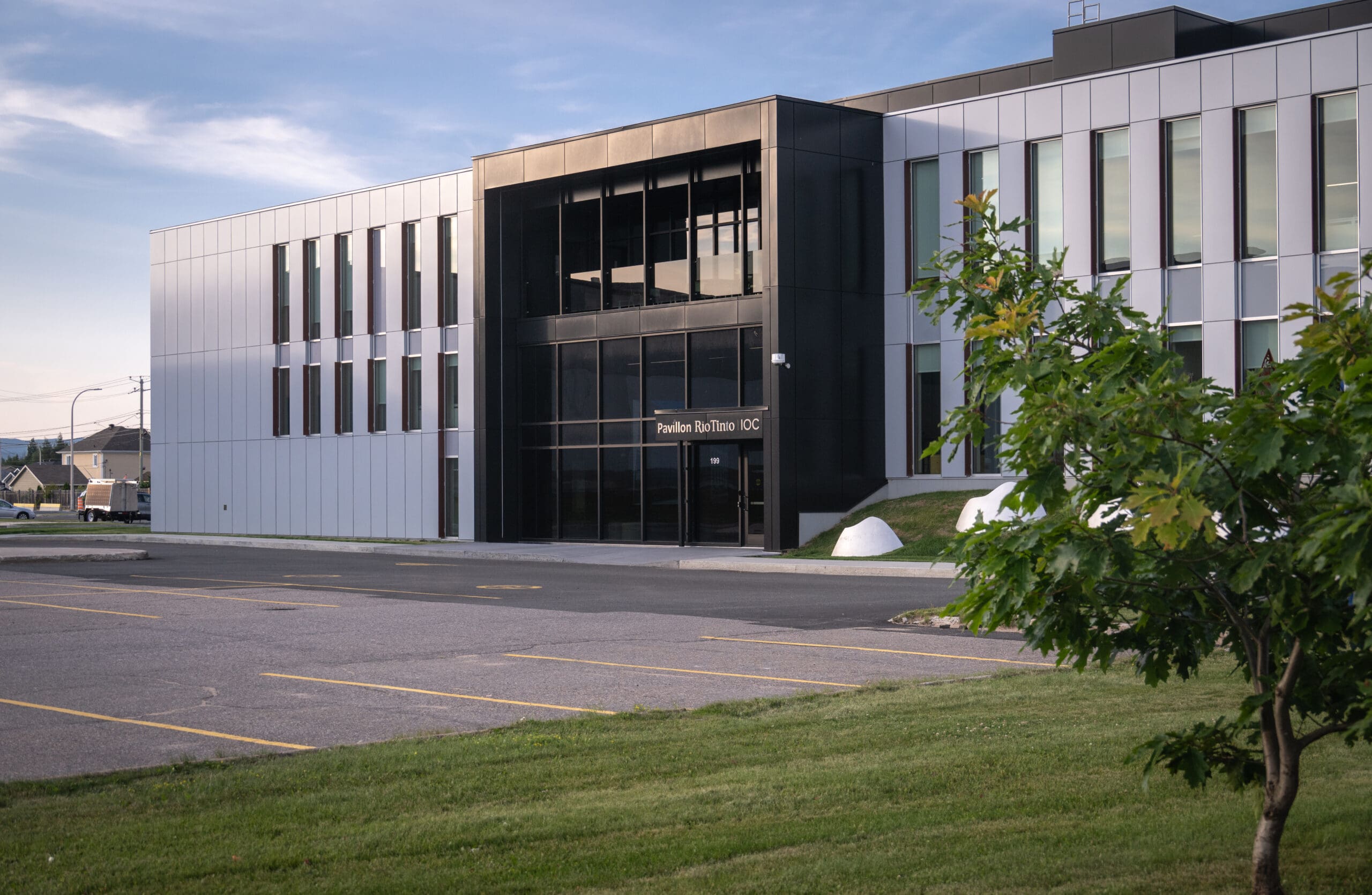 Immeuble de bureaux moderne doté de grandes baies vitrées, avec des panneaux extérieurs noirs et blancs, un parking vide et un petit arbre au premier plan, sous un ciel partiellement nuageux.