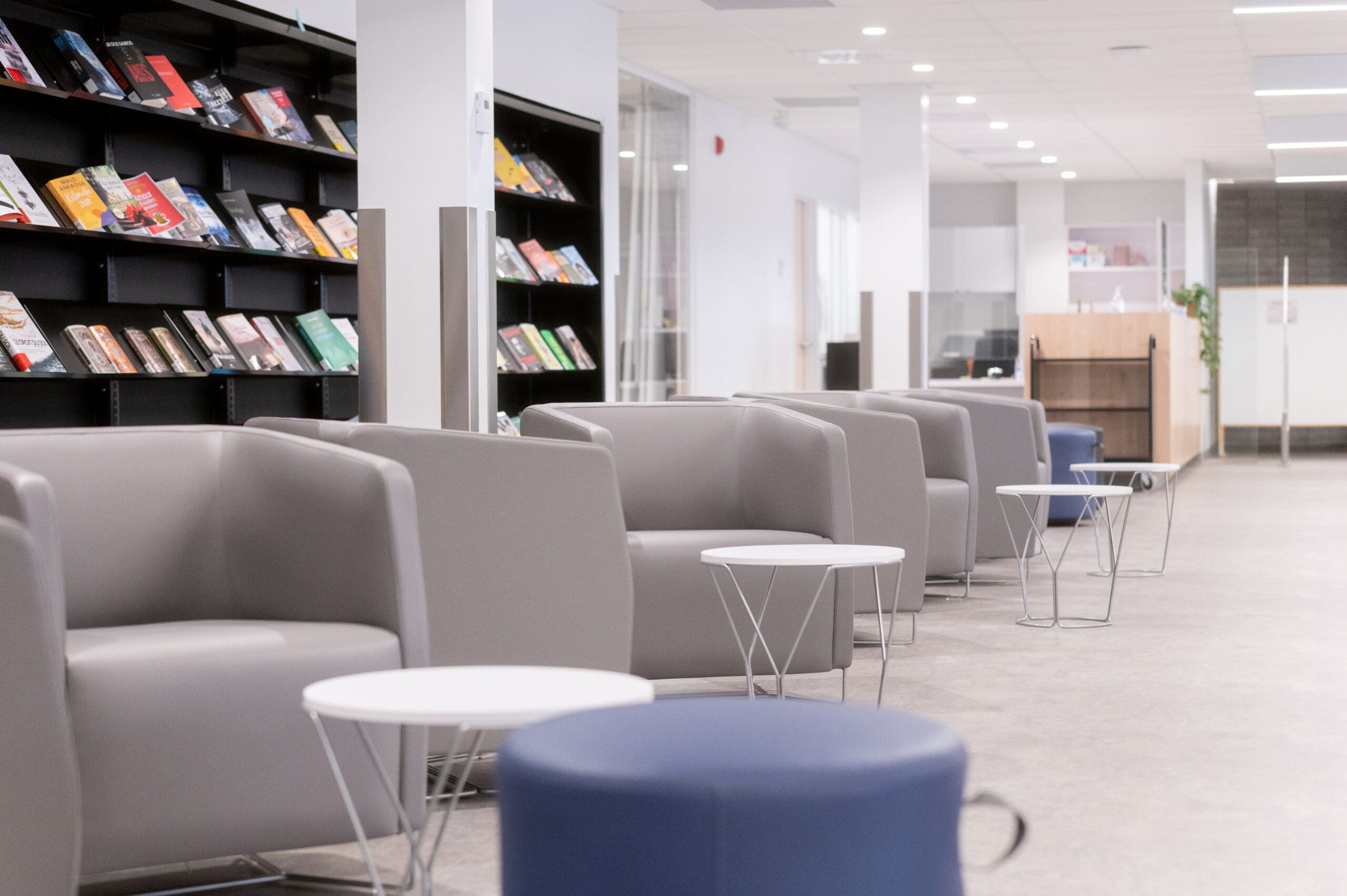Un salon-bibliothèque moderne et lumineux, avec des fauteuils gris, des petites tables rondes et des étagères remplies de livres colorés sur le mur de gauche. L'espace est ouvert et accueillant.