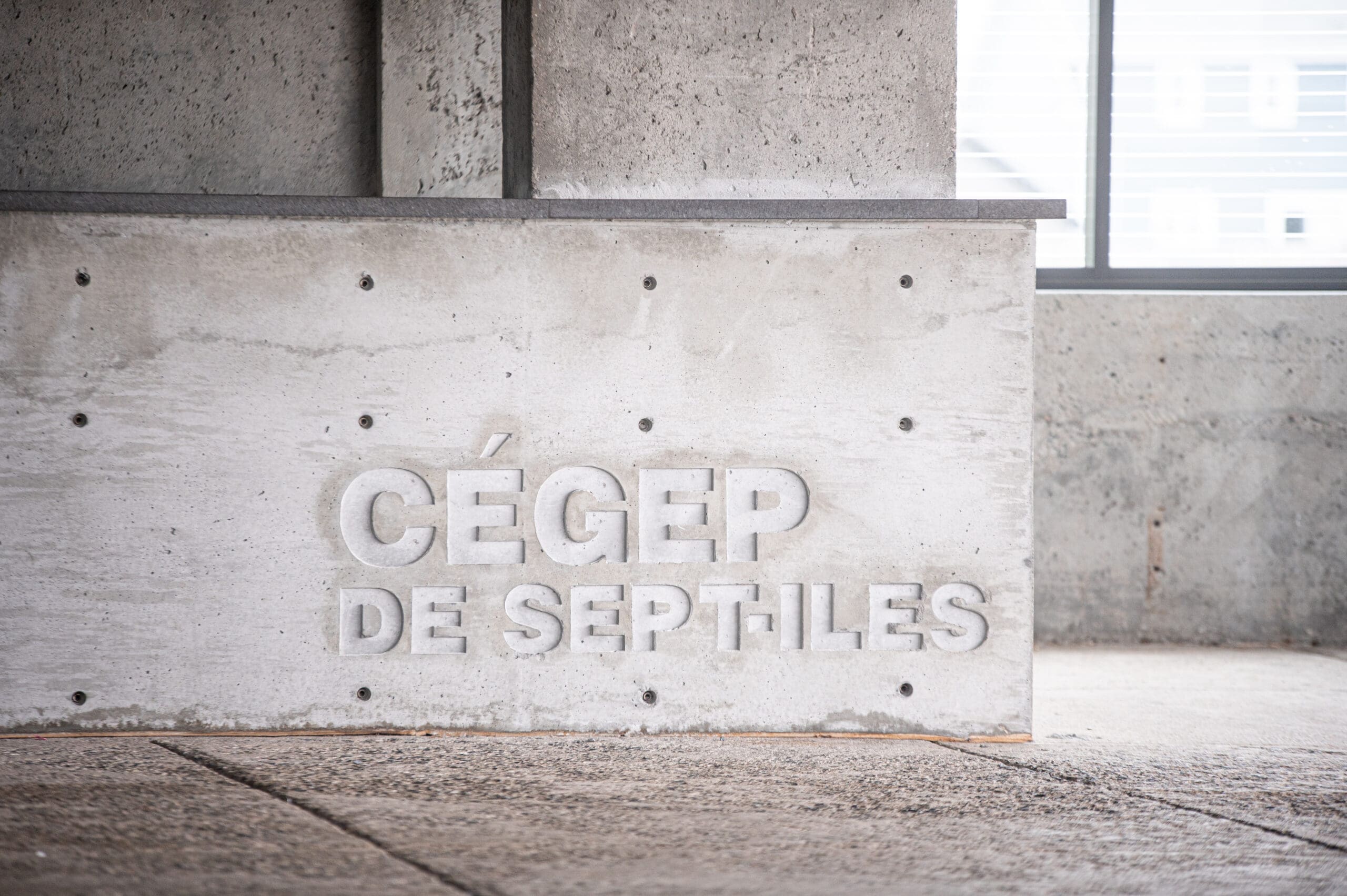 Mur en béton sur lequel est gravé le texte « CÉGEP DE SEPT-ÎLES » dans un bâtiment moderne, avec une fenêtre et de la lumière naturelle en arrière-plan.