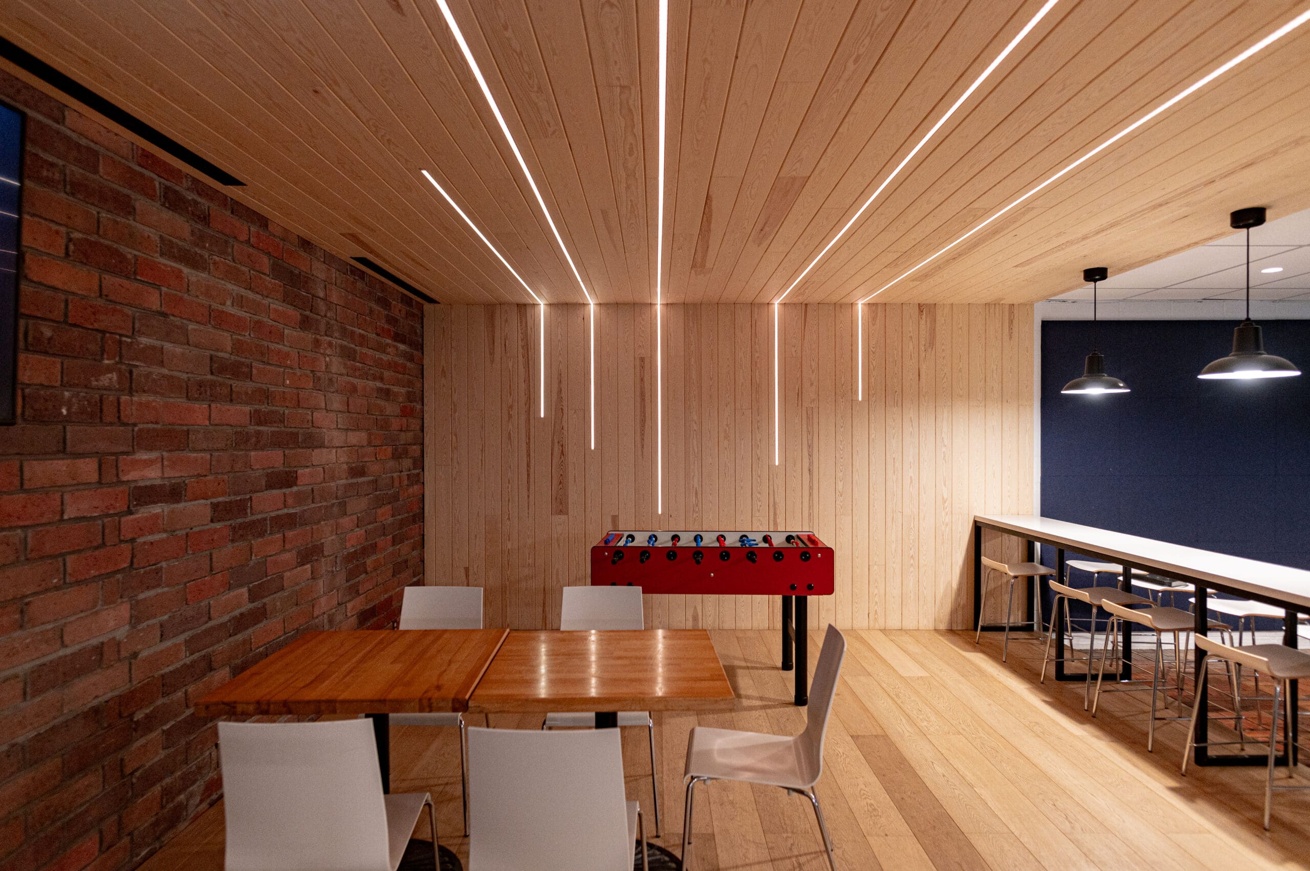 Salon moderne avec des murs et un parquet en bois, un baby-foot rouge, une table carrée entourée de chaises blanches, des tabourets de bar et des suspensions noires. Un mur d'accent en briques et des luminaires linéaires au plafond apportent une touche d'élégance.