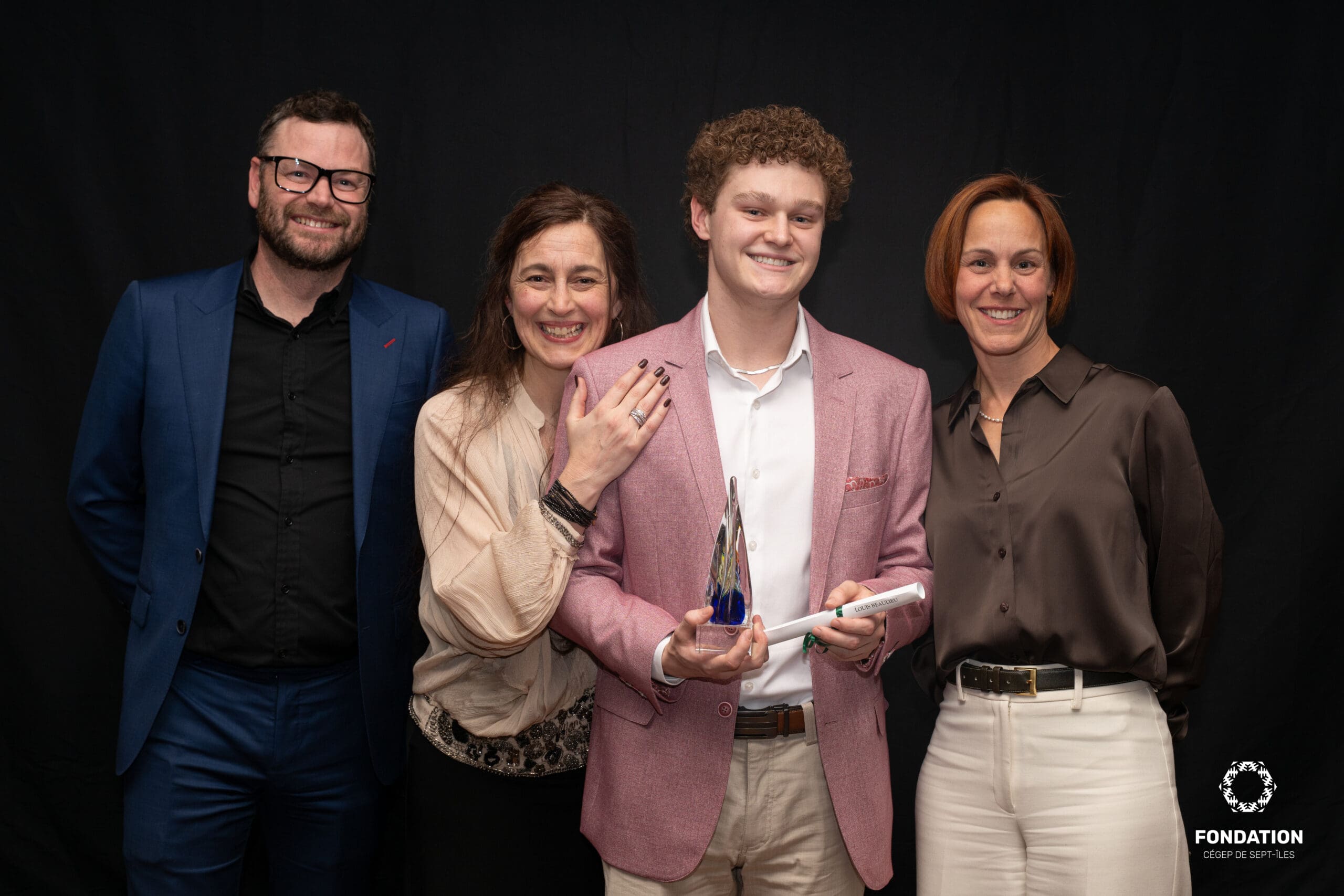 Quatre personnes posent en souriant sur un fond noir ; le jeune homme en veste rose tient un trophée et un diplôme, tandis que les autres se tiennent près de lui pour le soutenir. Un petit logo de la Fondation du Cégep de Sept-Îles figure dans le coin inférieur droit.