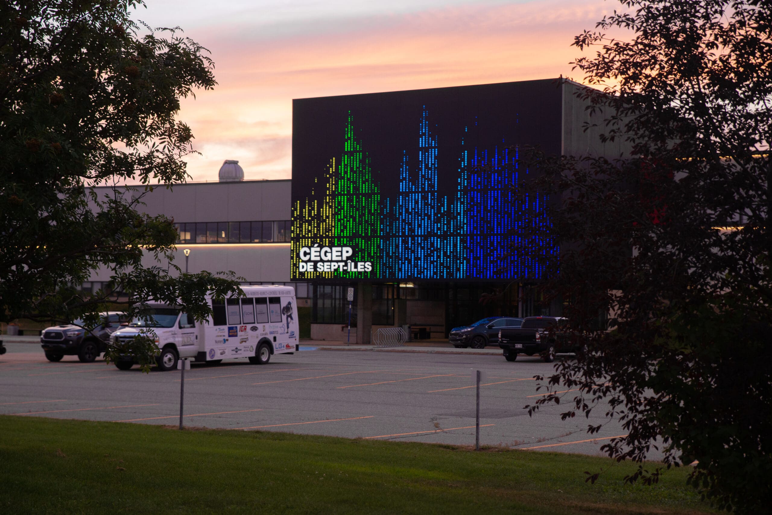 Au coucher du soleil, le bâtiment du Cégep de Sept-Îles arbore sur sa façade un grand écran numérique aux couleurs vives, avec quelques véhicules garés devant et des arbres qui encadrent partiellement la scène.