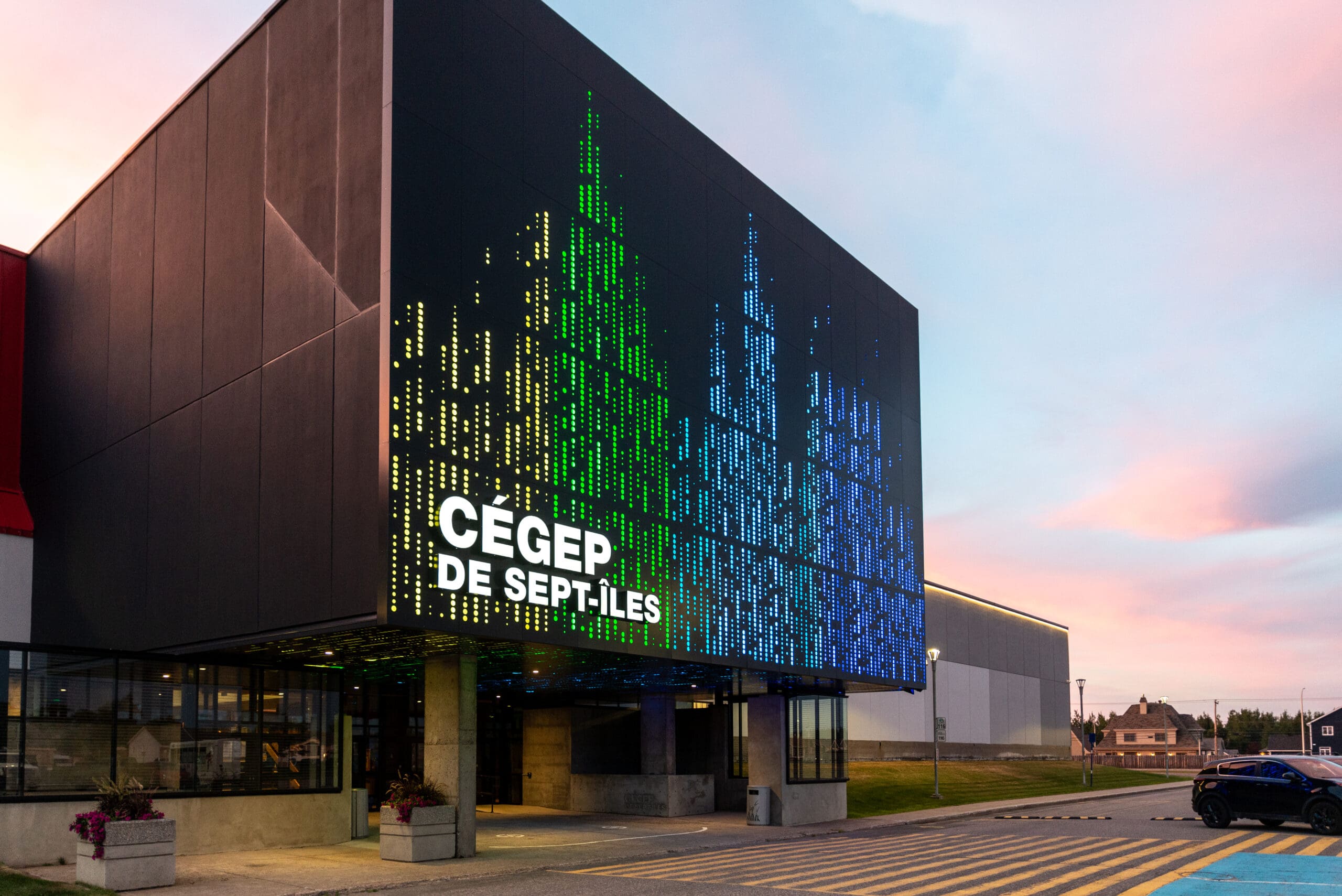 Bâtiment moderne dont la façade, à l'entrée, est ornée d'un écran numérique affichant des formes abstraites colorées et l'inscription « CÉGEP DE SEPT-ÎLES », avec un ciel au coucher du soleil en arrière-plan.