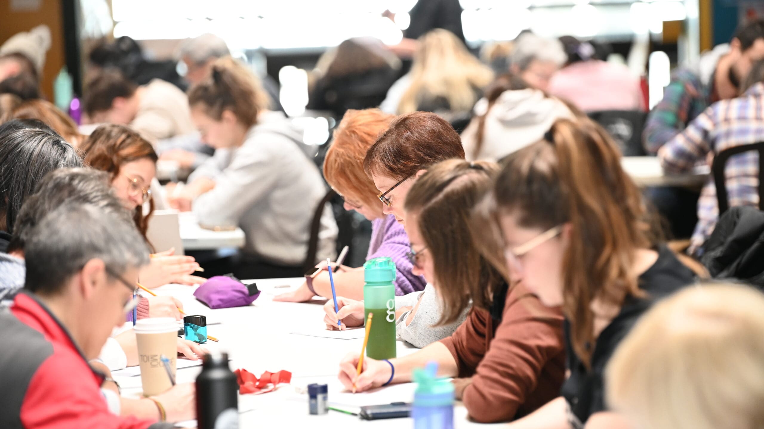 Un groupe de personnes est assis à des tables, les unes près des autres, concentrées sur leur travail ou leurs notes, avec des cahiers, du café et des bouteilles d'eau devant elles, dans un intérieur lumineux et animé.