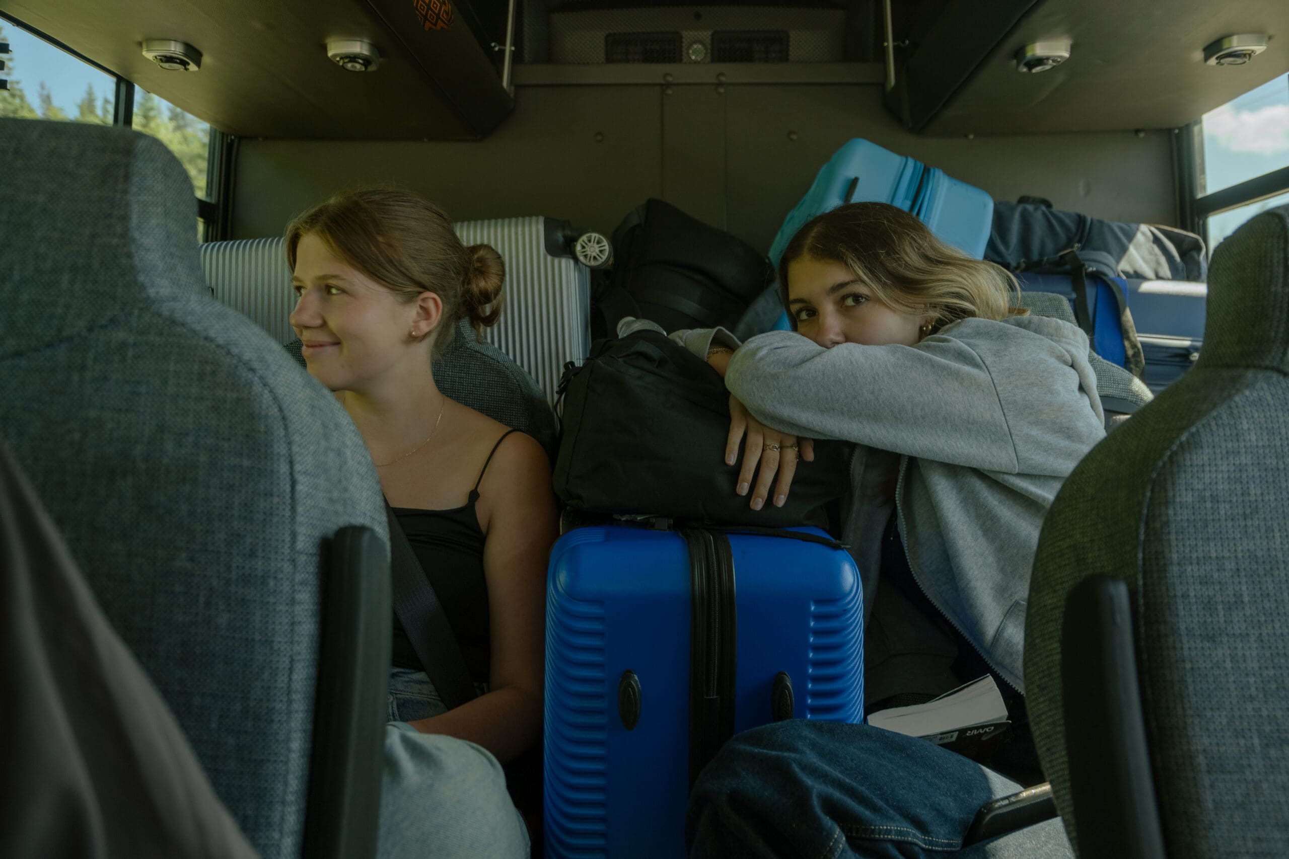 Deux jeunes sont assis dans un bus aux sièges gris, entourés de bagages, parmi lesquels une valise bleue bien visible. L'un d'eux sourit et regarde vers la gauche ; l'autre se penche en avant, les bras posés sur les sacs, et regarde vers la caméra.