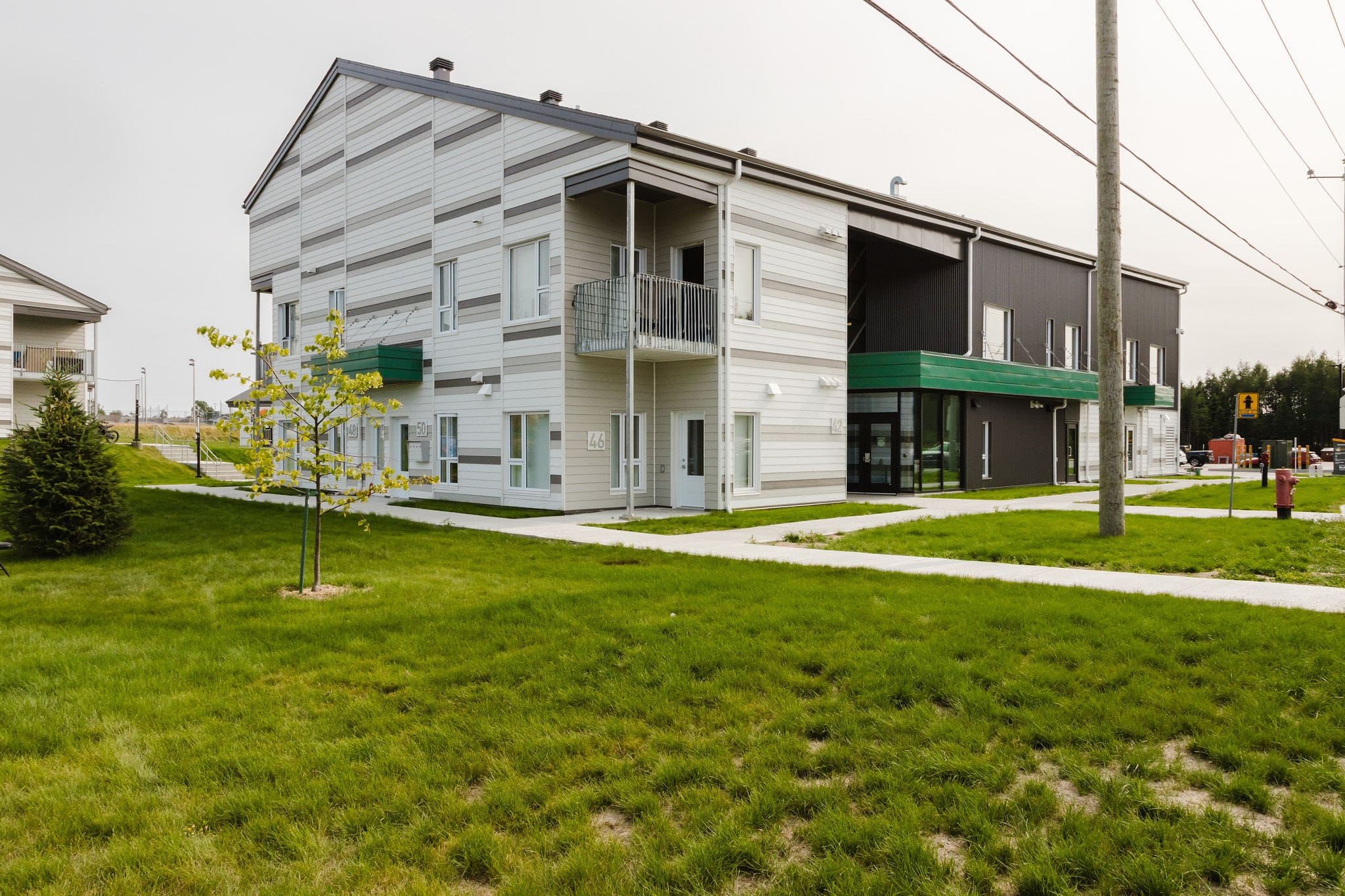 Un immeuble d'appartements moderne de deux étages, avec un bardage gris et blanc, des touches de vert, des balcons et de grandes fenêtres, entouré d'une pelouse et de trottoirs sous un ciel couvert.