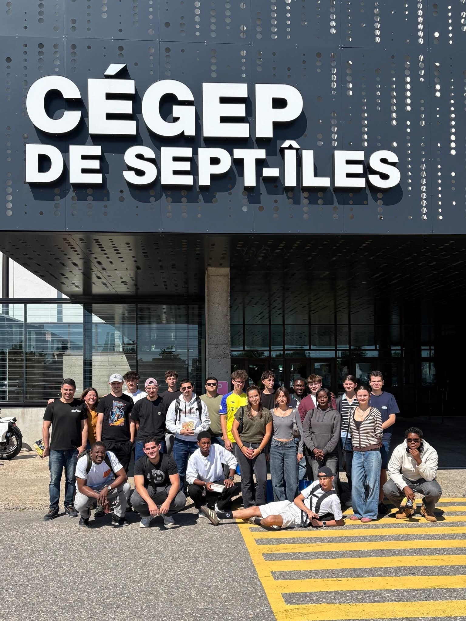 Un groupe d'environ 25 jeunes adultes posent ensemble à l'extérieur, sous un grand panneau sur lequel on peut lire « CÉGEP DE SEPT-ÎLES ». Le groupe se tient debout et à genoux près d'un passage piéton, par une journée ensoleillée.