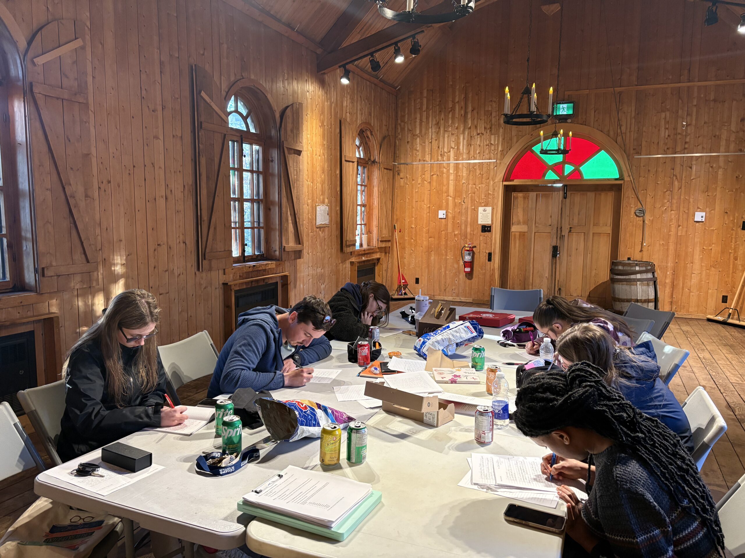 Six personnes sont assises autour d'une table dans une pièce aux murs en bois et écrivent sur des feuilles. Des en-cas, des boissons et des sacs sont posés sur la table. La pièce est dotée de grandes fenêtres, d'un tonneau et, au fond, d'une porte en vitrail.