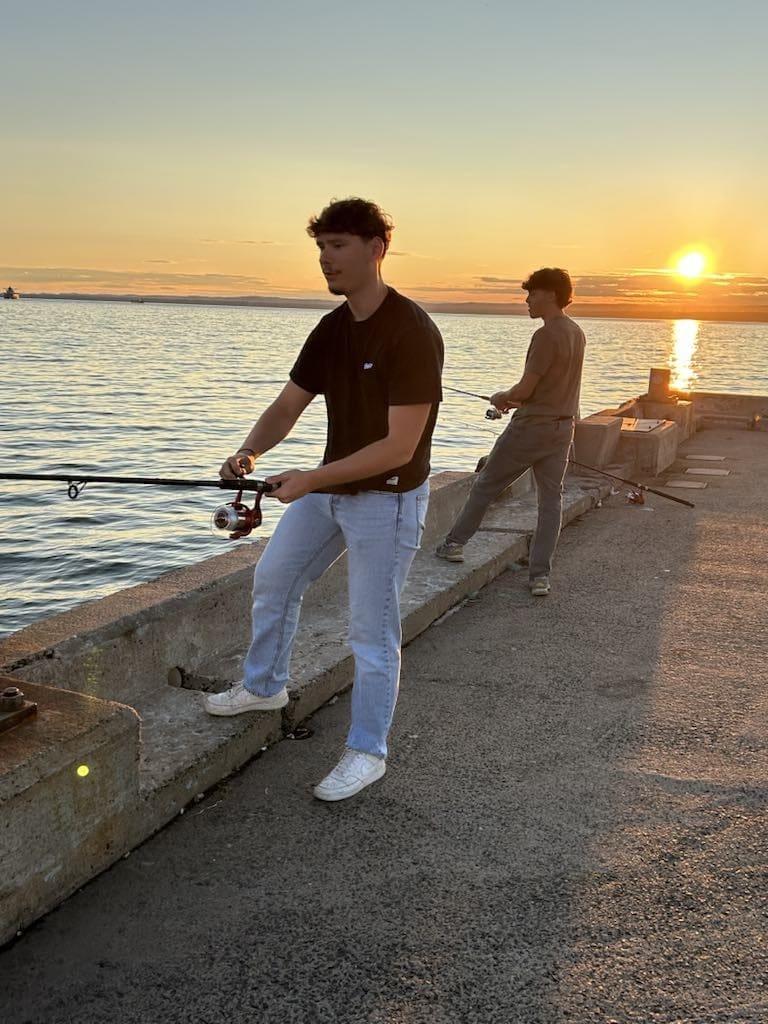 Deux personnes pêchent sur une jetée en béton au coucher du soleil. Le soleil est bas au-dessus de l'eau calme, projetant une lueur chaleureuse. L'une des deux personnes est plus proche de l'appareil photo, une canne à pêche à la main, tandis que l'autre se tient un peu plus en retrait.