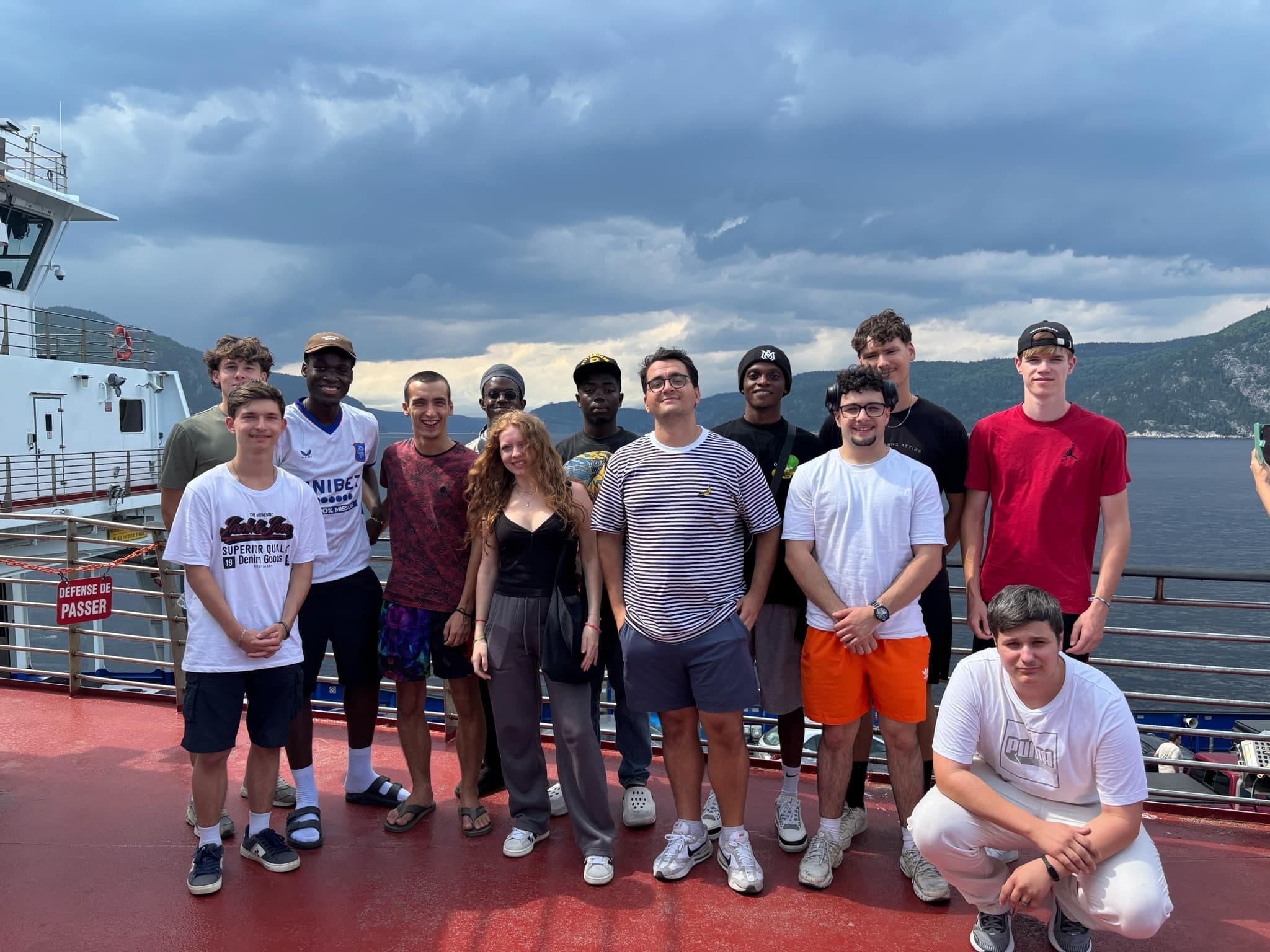 Un groupe de treize jeunes prend la pose et sourit sur le pont d'un ferry, avec les montagnes et l'eau en arrière-plan, sous un ciel nuageux.