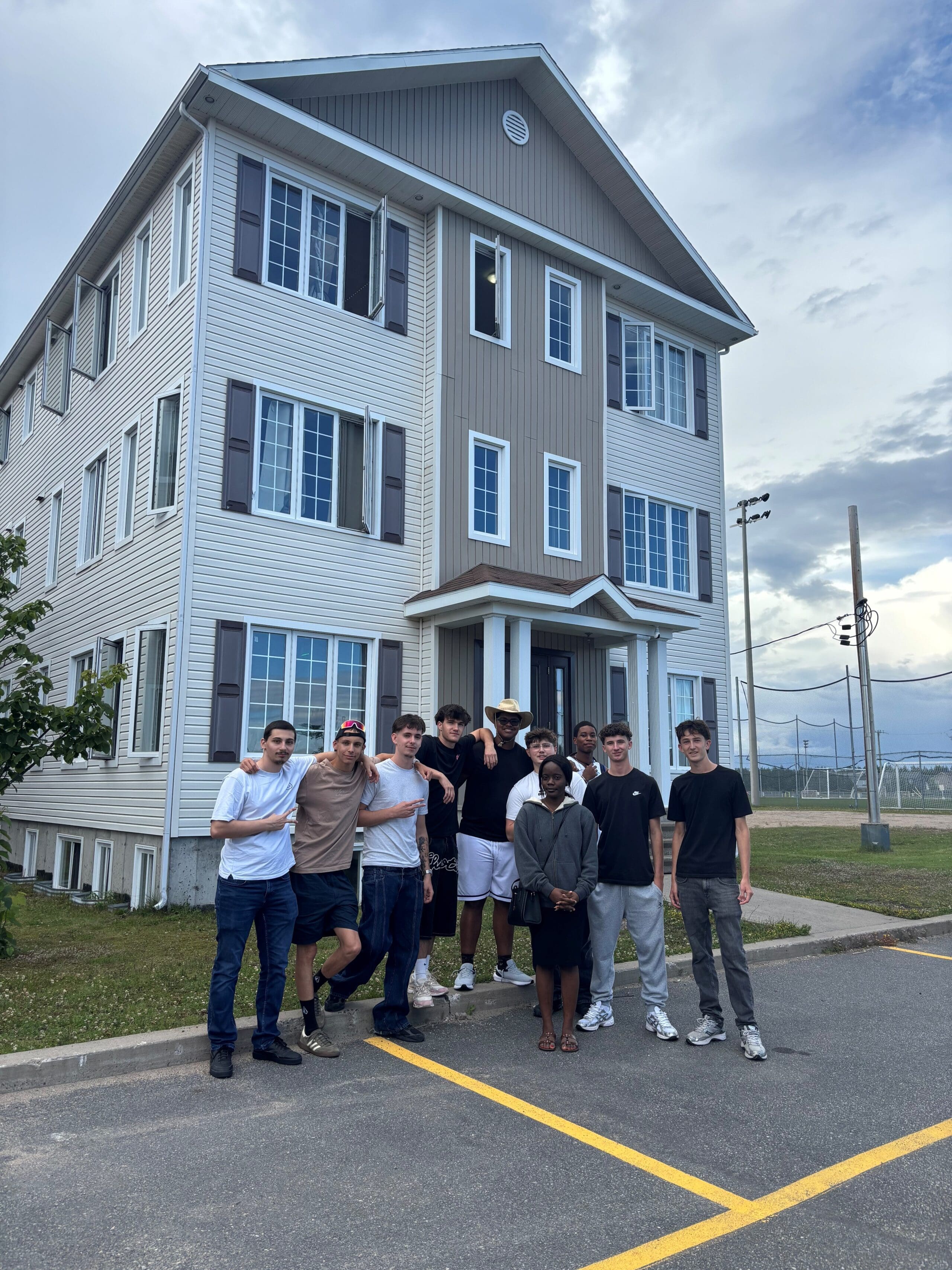 Neuf personnes se tiennent côte à côte et posent pour une photo de groupe devant un immeuble de trois étages aux volets gris, par une journée nuageuse, près d'un parking et de terrains de sport.