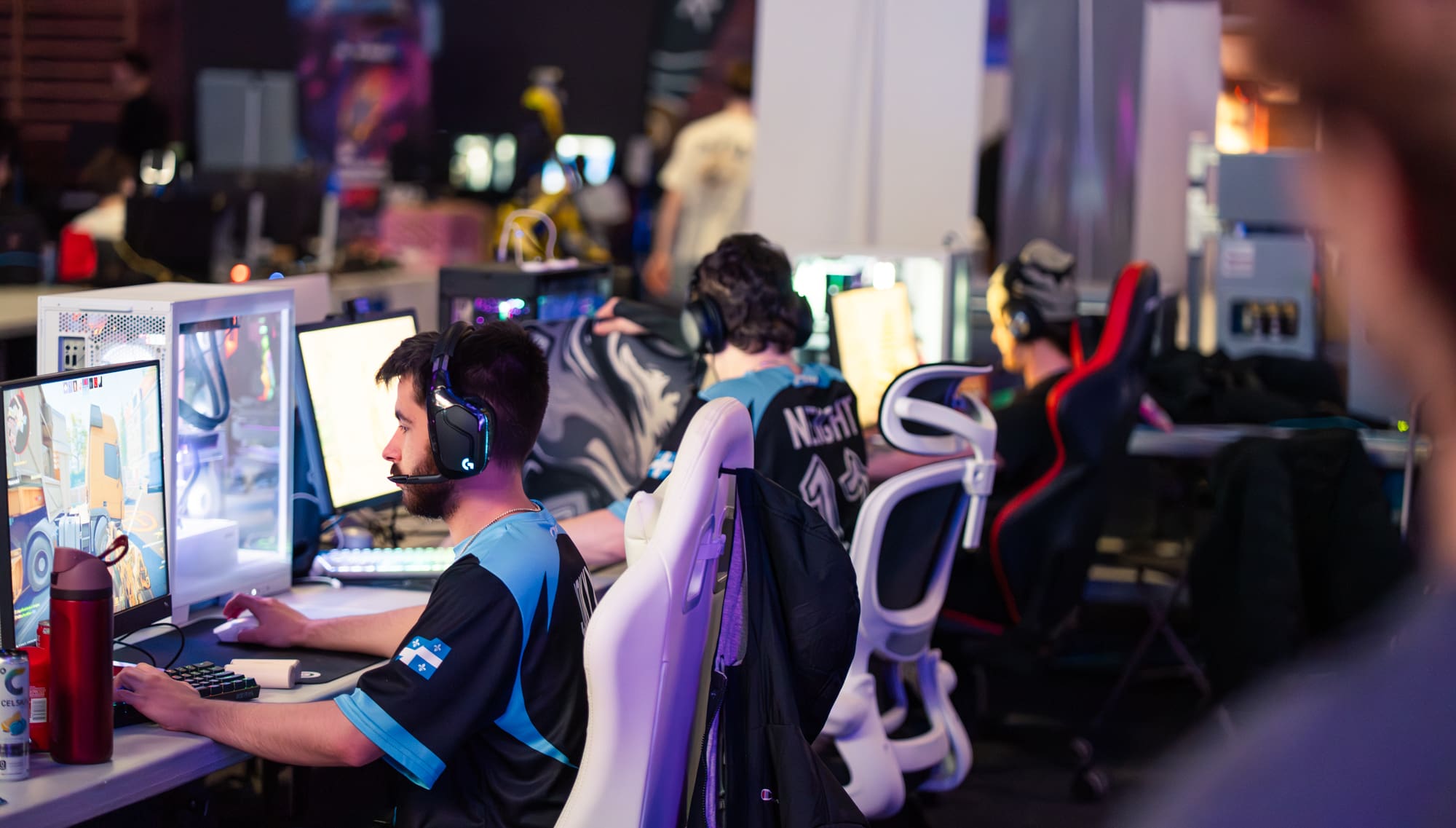 Lors d'un événement de gaming très éclairé, plusieurs joueurs équipés de casques s'affrontent devant des ordinateurs de bureau. Assis dans des fauteuils ergonomiques, ils sont concentrés sur leurs écrans, leur matériel de gaming et leurs bouteilles d'eau posés sur leurs bureaux.