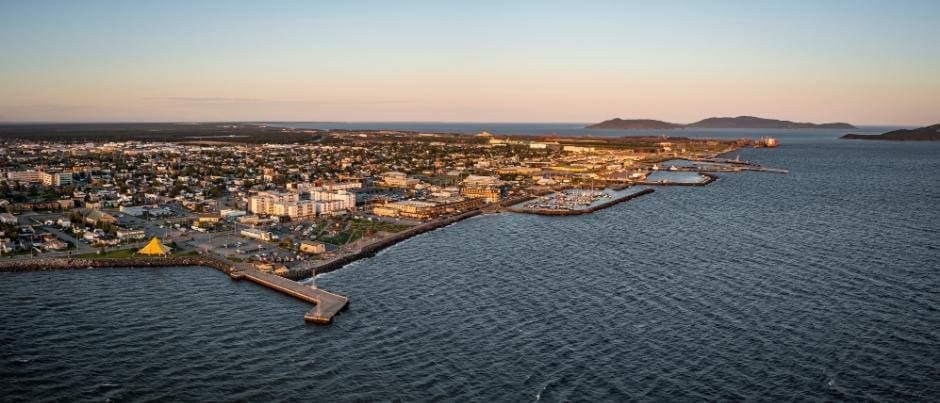Vue aérienne d'une ville côtière avec ses bâtiments, ses jetées et ses quais s'avançant dans une vaste étendue d'eau au coucher du soleil, avec des collines lointaines à l'horizon sous un ciel dégagé.