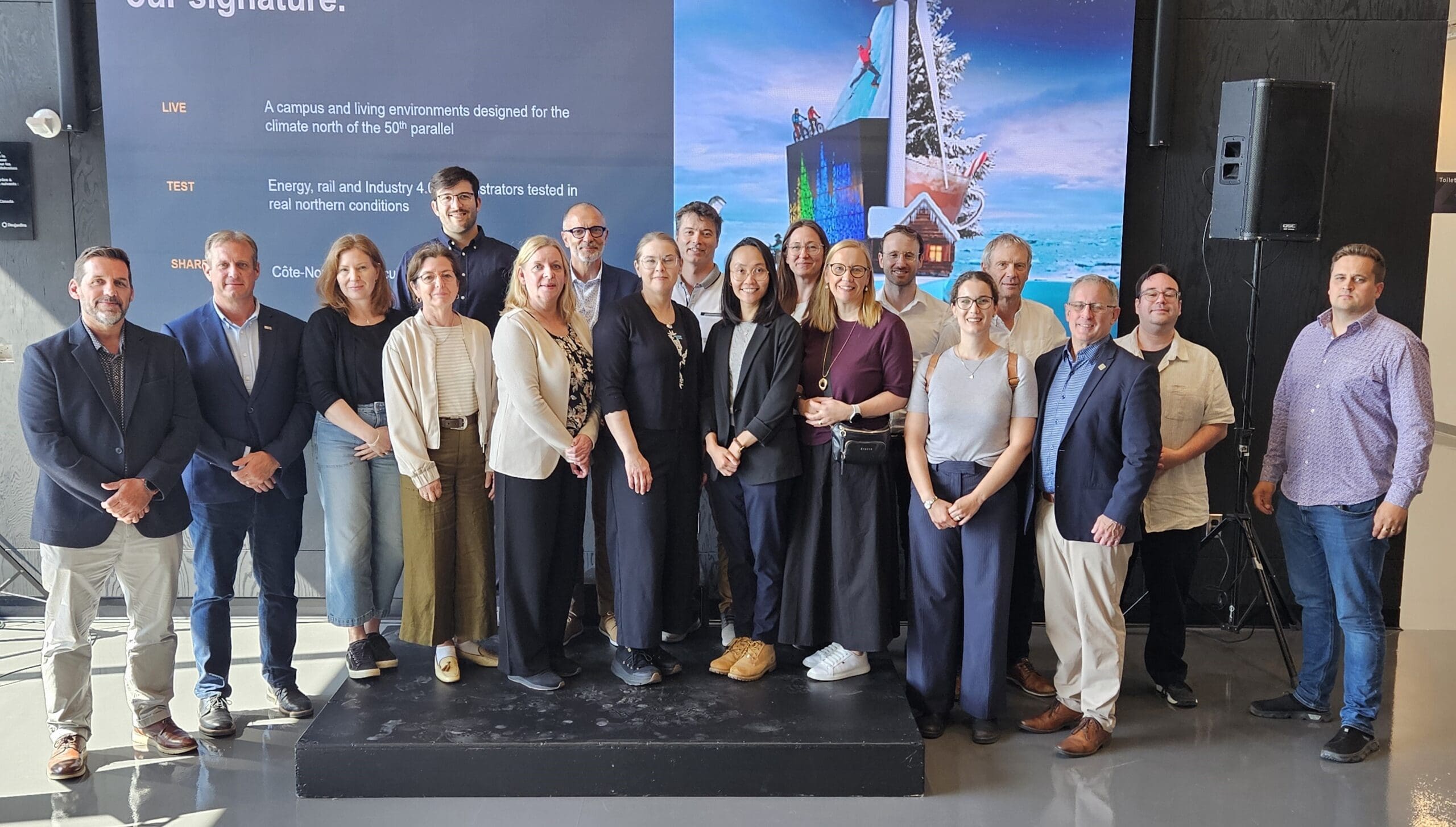Un groupe de personnes pose pour une photo à l'intérieur, devant des panneaux d'affichage, dont certains comportent du texte et des images de bâtiments enneigés. La plupart sourient et portent une tenue professionnelle décontractée.