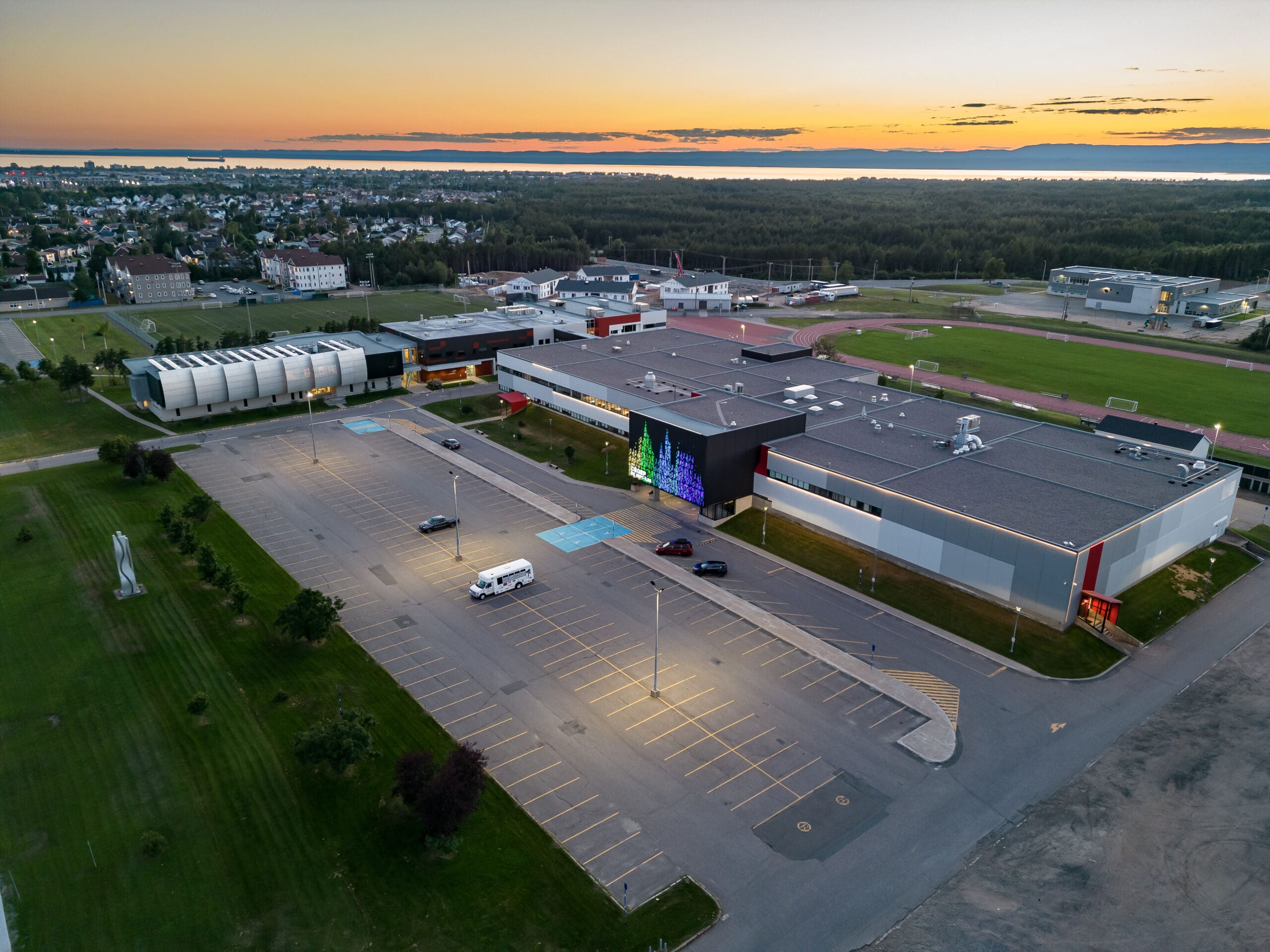 Vue aérienne d'un grand bâtiment moderne avec un parking presque vide au coucher du soleil. Le bâtiment est doté d'un écran numérique coloré sur l'un de ses murs, entouré de champs verdoyants et d'un quartier résidentiel en arrière-plan.