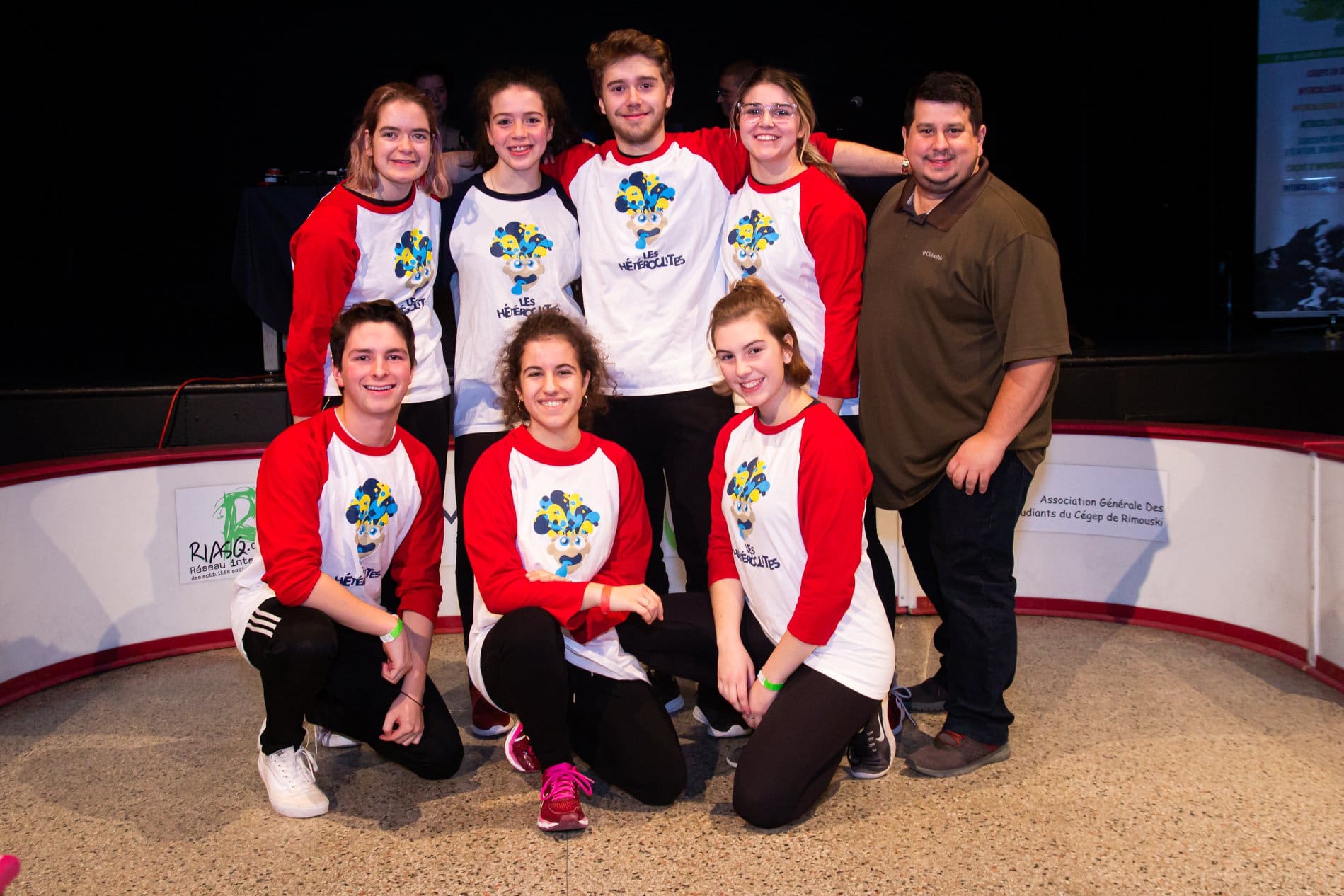 Un groupe de sept jeunes, vêtus de maillots rouges et blancs assortis, posent et sourient aux côtés d'un homme en maillot vert, sur une patinoire couverte avec un fond noir.