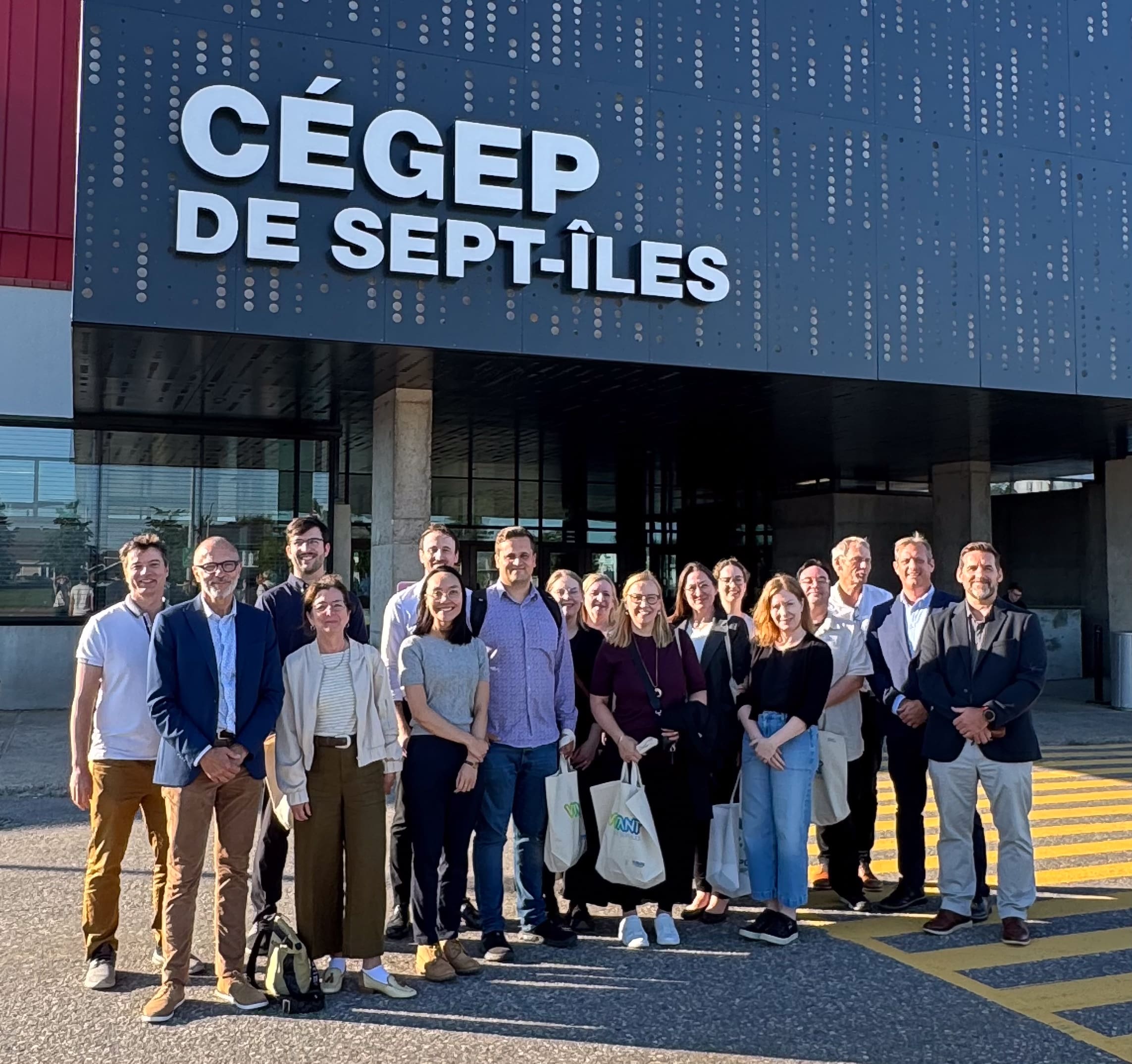 Un groupe de personnes se tient souriante devant le bâtiment du Cégep de Sept-Îles par une journée ensoleillée. Certaines tiennent des sacs et le groupe semble prendre la pose pour une photo de groupe près de l'entrée.